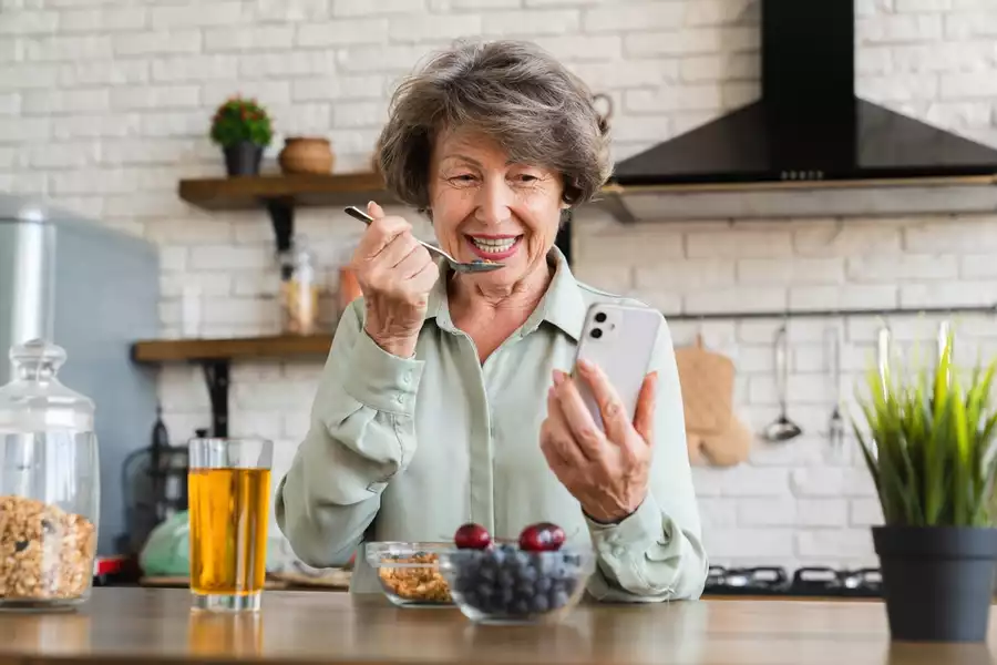Cheerful elderly woman eating a bowl of fruit and granola in the kitchen
